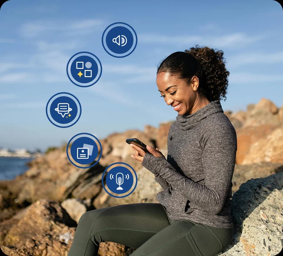 A woman sitting outdoors on rocks near water, smiling and looking at her phone, with Rosetta Stone Sapphire feature icons overlaid around her.